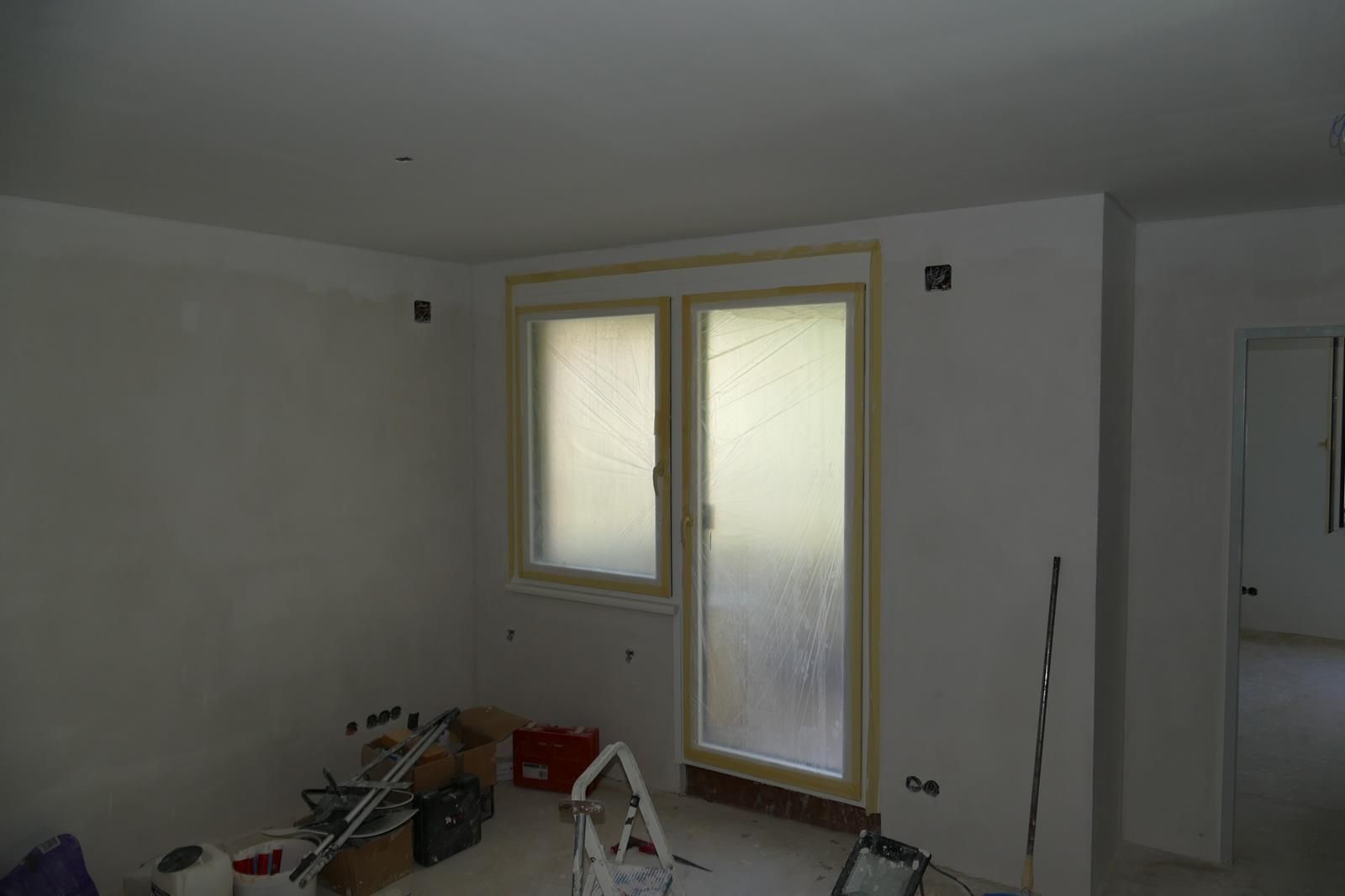 Ein Innenraum im Umbauprozess mit kahlen weißen Wänden, einem Fenster, einer Balkontür und Bauwerkzeugen auf dem Boden.