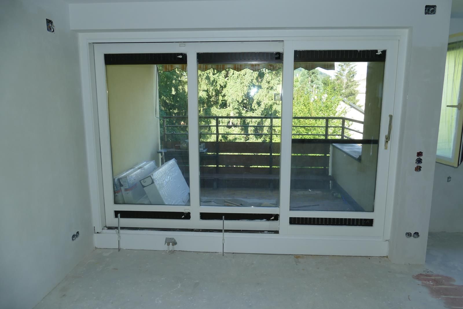 Große weiße Glasschiebetüren wurden in einem im Bau befindlichen Raum installiert und bieten einen Blick auf einen Balkon und die umliegenden Bäume.