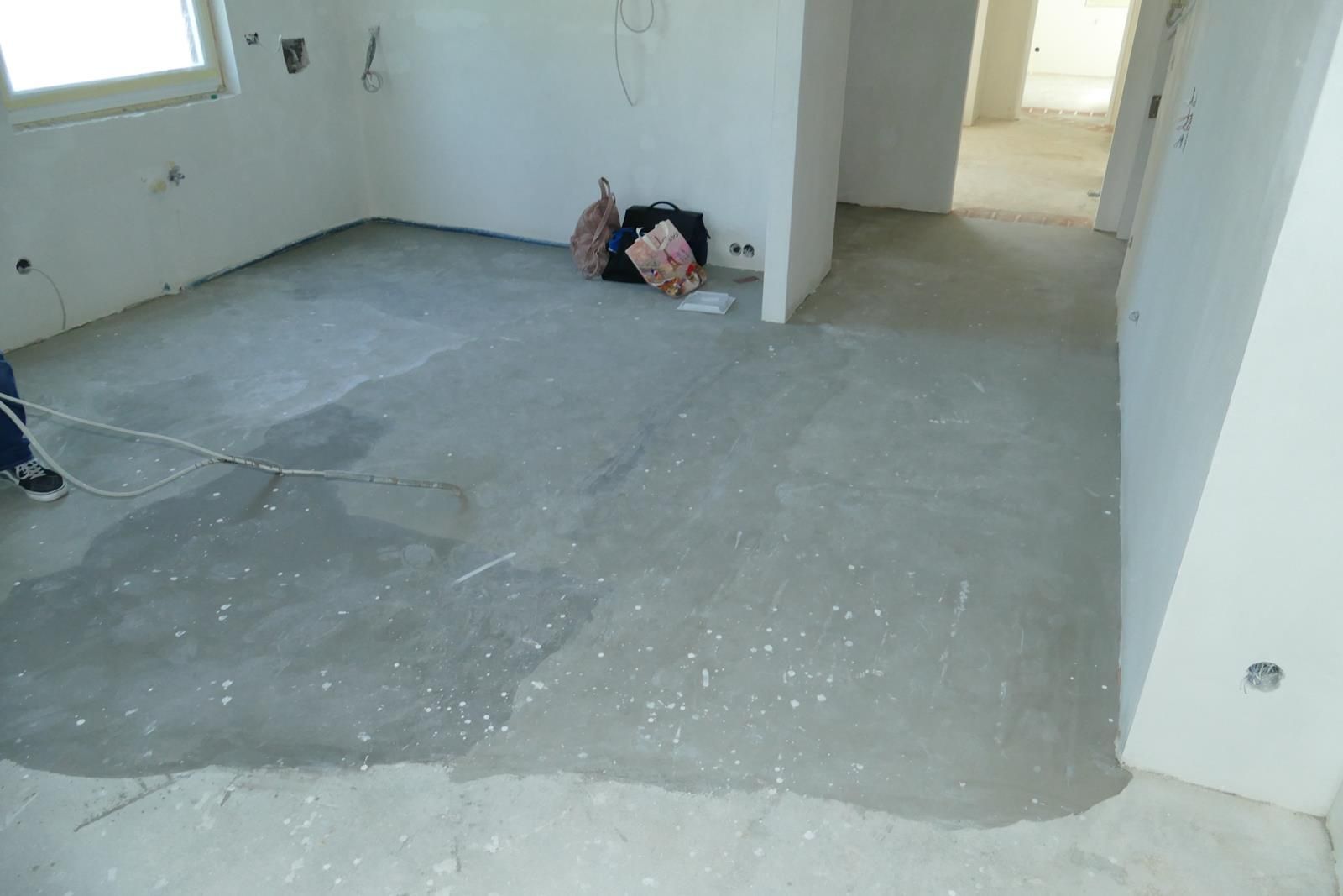 Eine Person sitzt auf dem Boden eines Zimmers mit unfertigem Betonboden und weißen Wänden, vermutlich während einer Hausrenovierung.