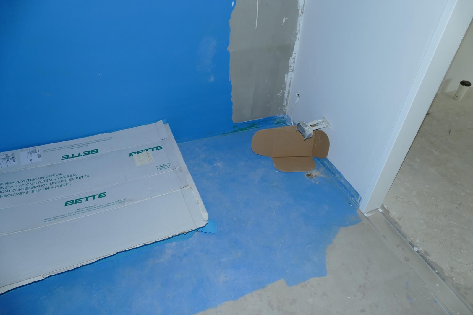 Eine Baustellenecke mit blauer Bodenabdichtung, weißen Tafeln und freiliegenden Wasserleitungen an einer Wand.