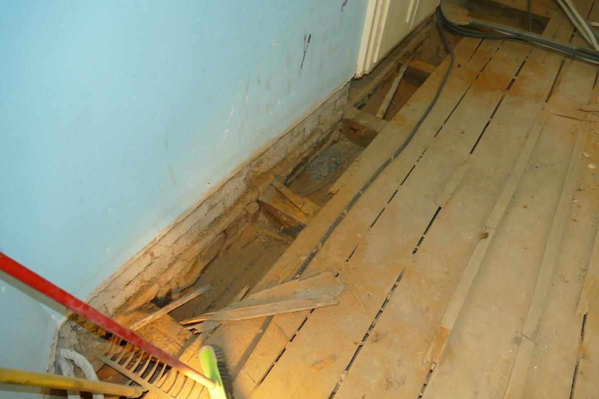 Ein teilweise entfernter Holzboden gibt den Blick auf die Balken und einen dunklen Spalt entlang einer hellblauen Wand frei, was auf Renovierungsarbeiten im Haus hindeutet.