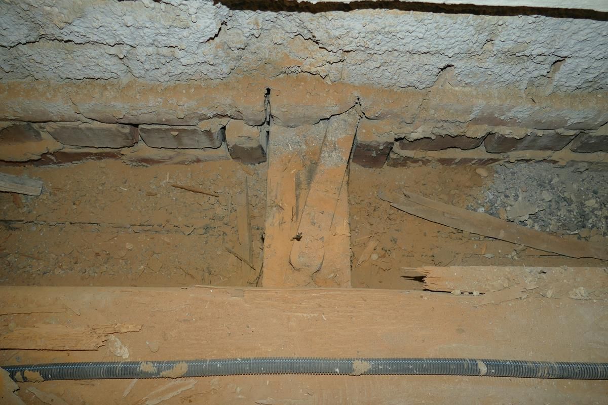 Eine Nahaufnahme einer Baustelle zeigt ein Betonfundament, das mit einer Ziegelmauer verbunden ist, wobei die Bewehrungsstäbe im Sand freiliegen.