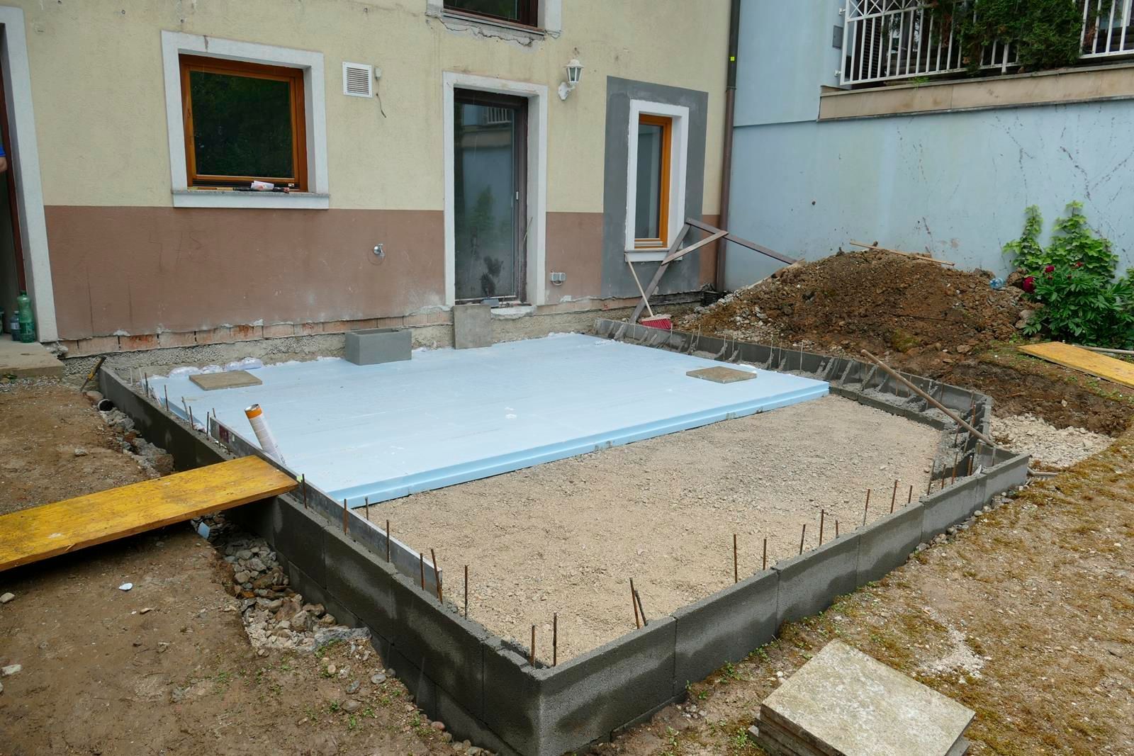 Eine Baustelle, die einen mit Kies gefüllten Betonfundamentumfang und einen mit blauen Schaumstoffplatten abgedeckten Bereich zeigt.