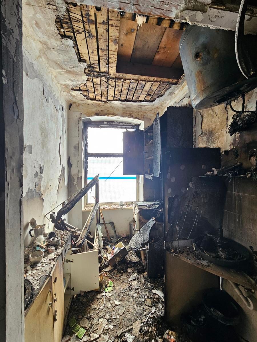 Ein kleiner, durch einen Brand beschädigter Raum mit freiliegenden Holzbalken an der Decke, mit Trümmern bedeckten Böden und einem hellen Fenster auf der Rückseite.