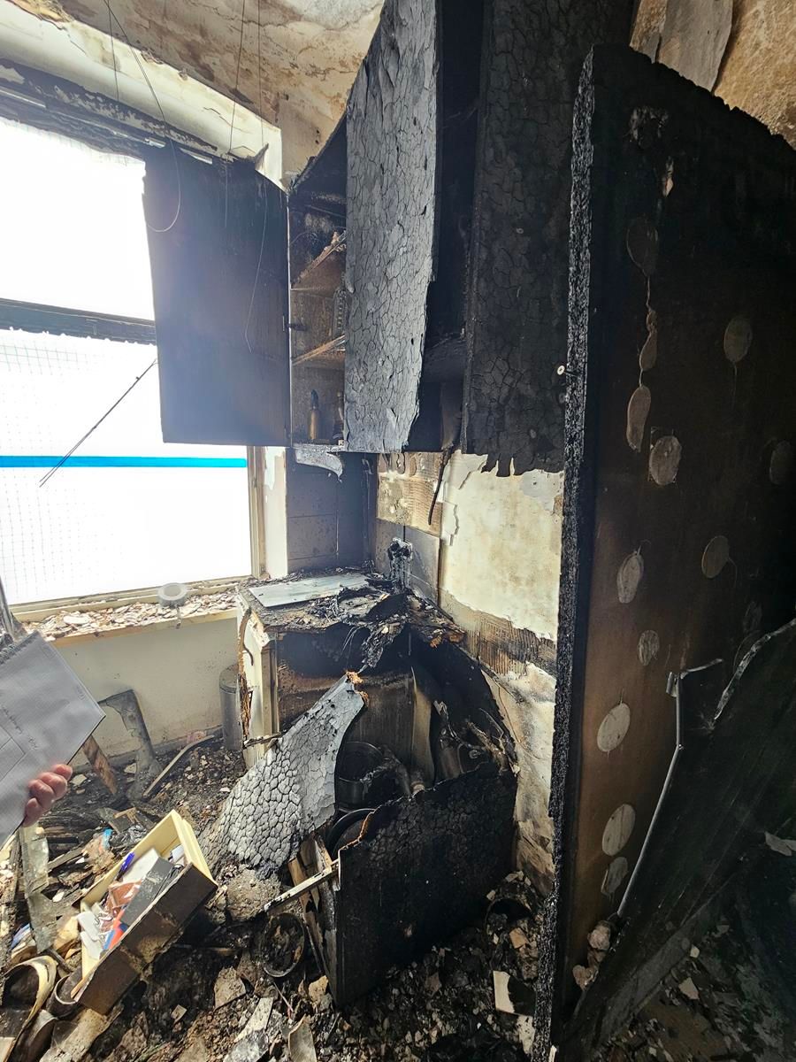 Ein stark durch einen Brand beschädigtes Kücheninterieur mit geschwärzten Schränken und Trümmern, gesehen neben einem Fenster.