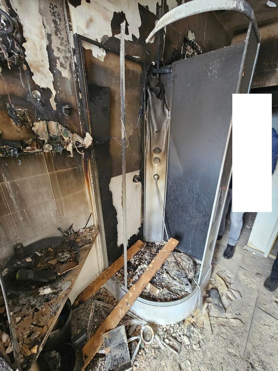 Eine durch ein Feuer stark beschädigte Duschkabine mit verkohlten Wänden und Trümmern in einem ansonsten beschädigten Raum.