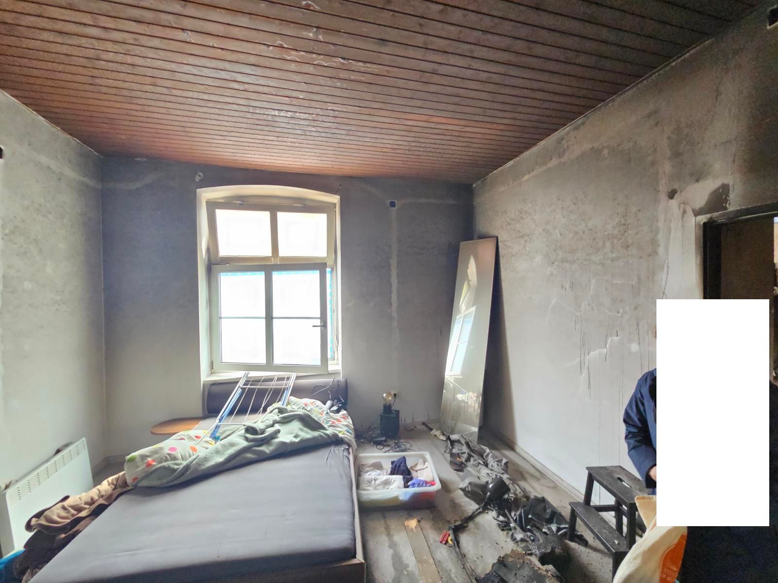 Ein spärlich möbliertes, unfertiges Zimmer mit einer Matratze am Fenster, einer Holzdecke und Betonwänden.