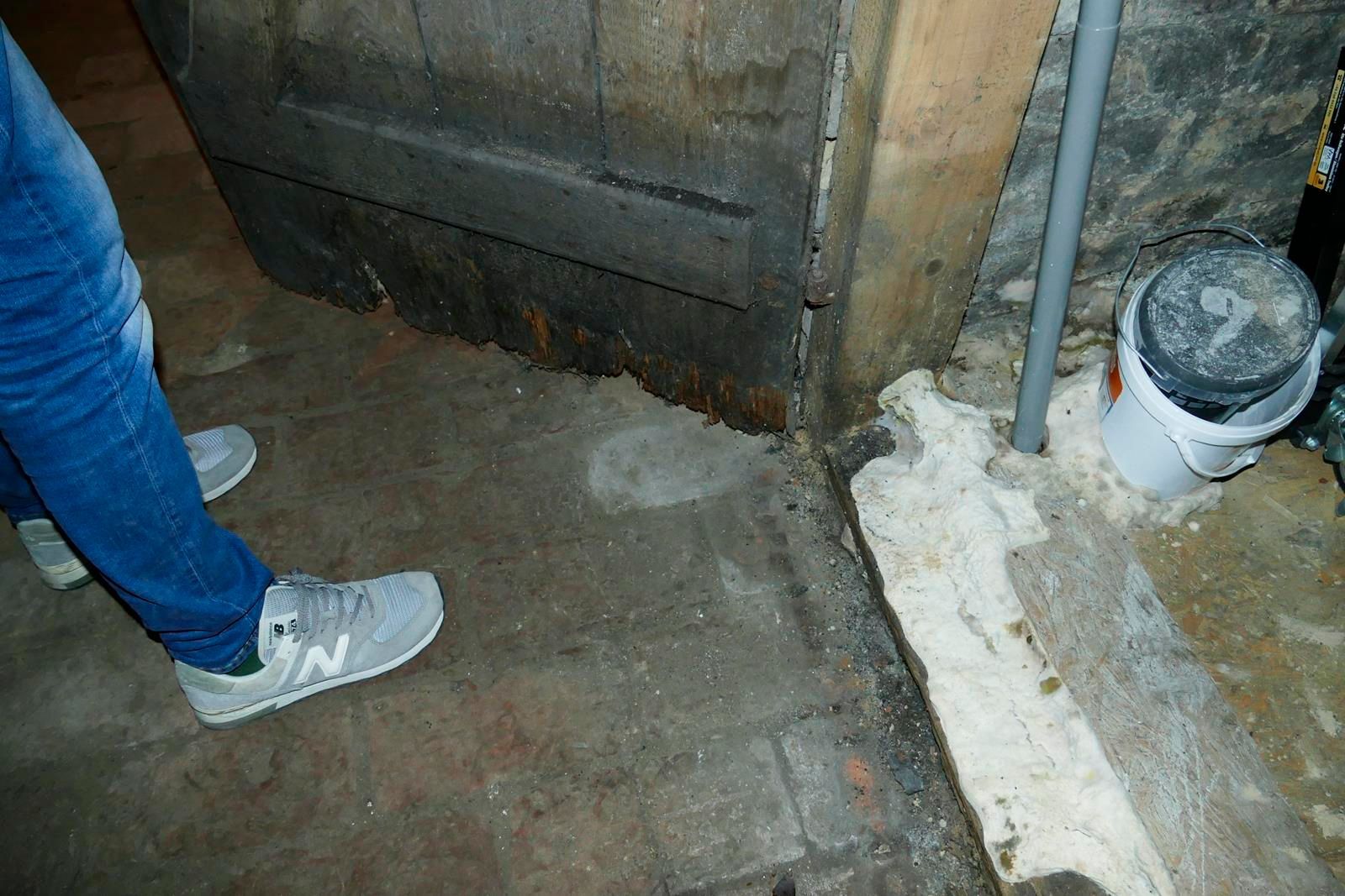 Eine Person in blauen Jeans und grauen Turnschuhen steht auf einem Ziegelboden neben einer abgenutzten Holztür und einem Plastikeimer.
