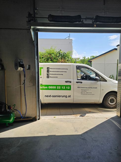 Ein weißer Lieferwagen mit grüner Aufschrift parkt vor einer Garage, aufgenommen aus dem abgedunkelten Arbeitsbereich.