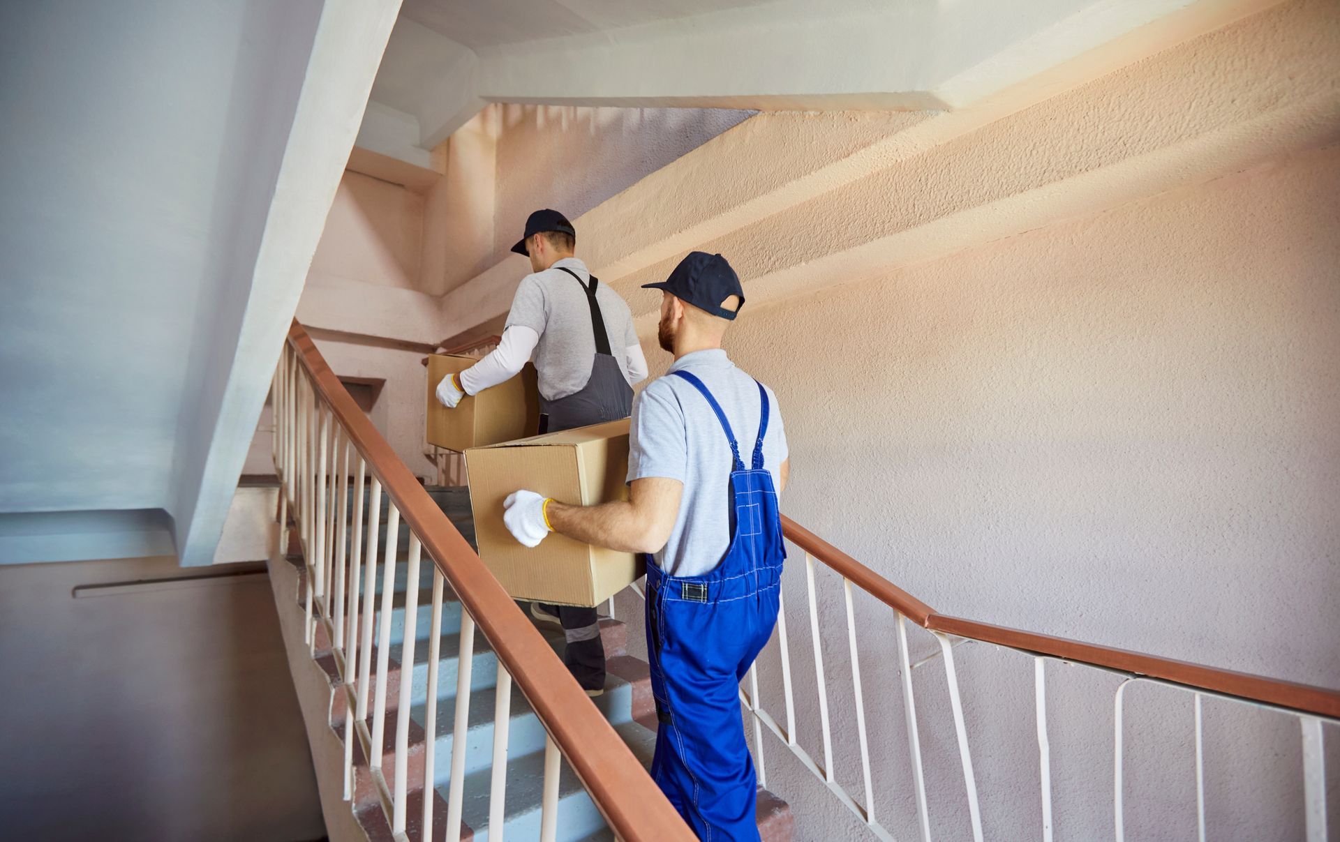 Two Movers in Blue Overalls and Caps Carrying Boxes Up a Staircase — Dan's Removals In Lismore, NSW