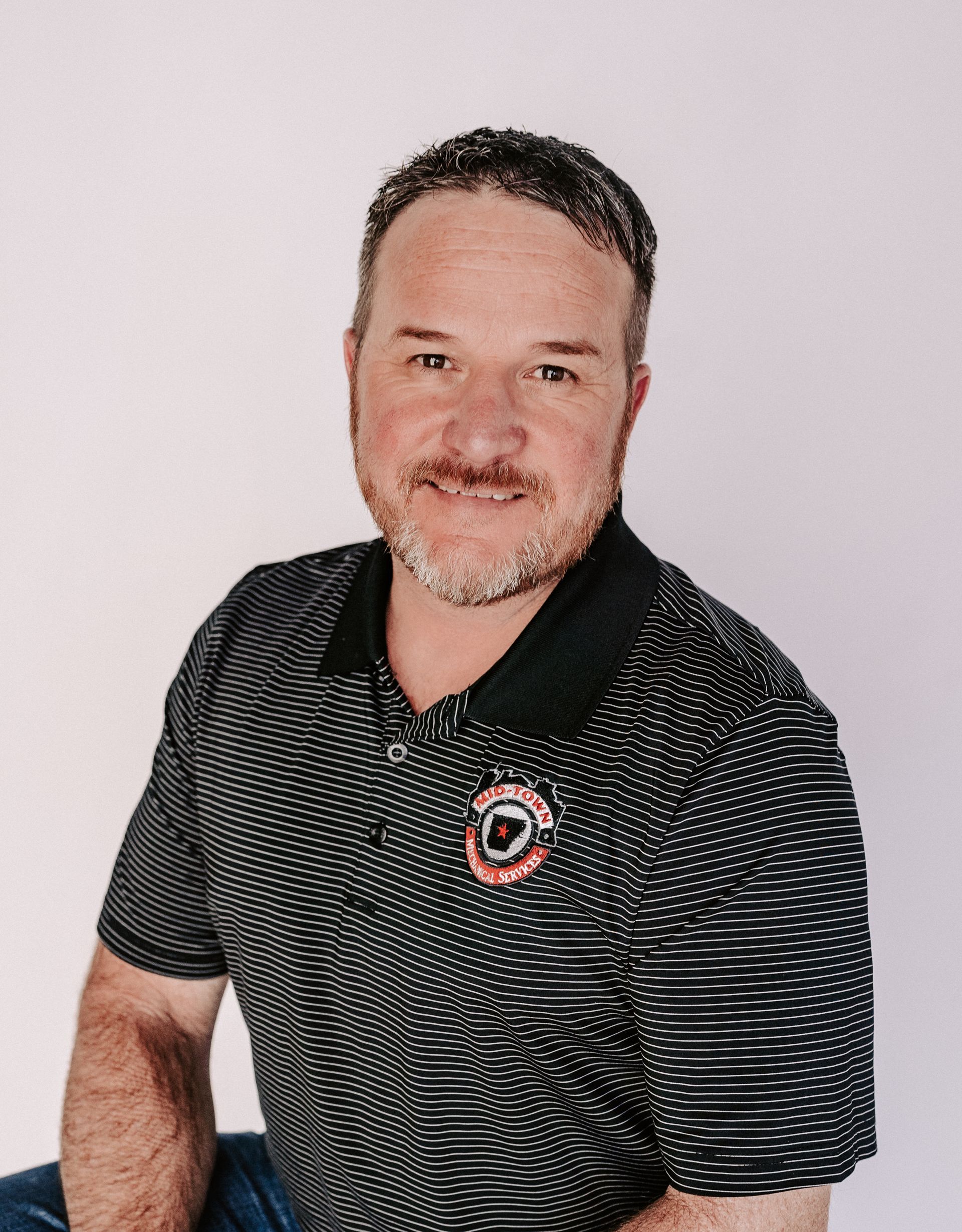 Man in a black striped polo shirt smiling against a light gray background.
