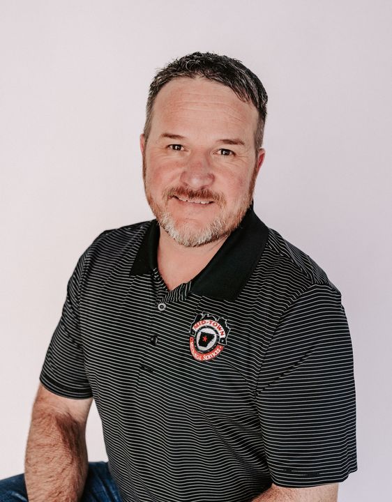 Man in a black patterned polo shirt smiling against a light background