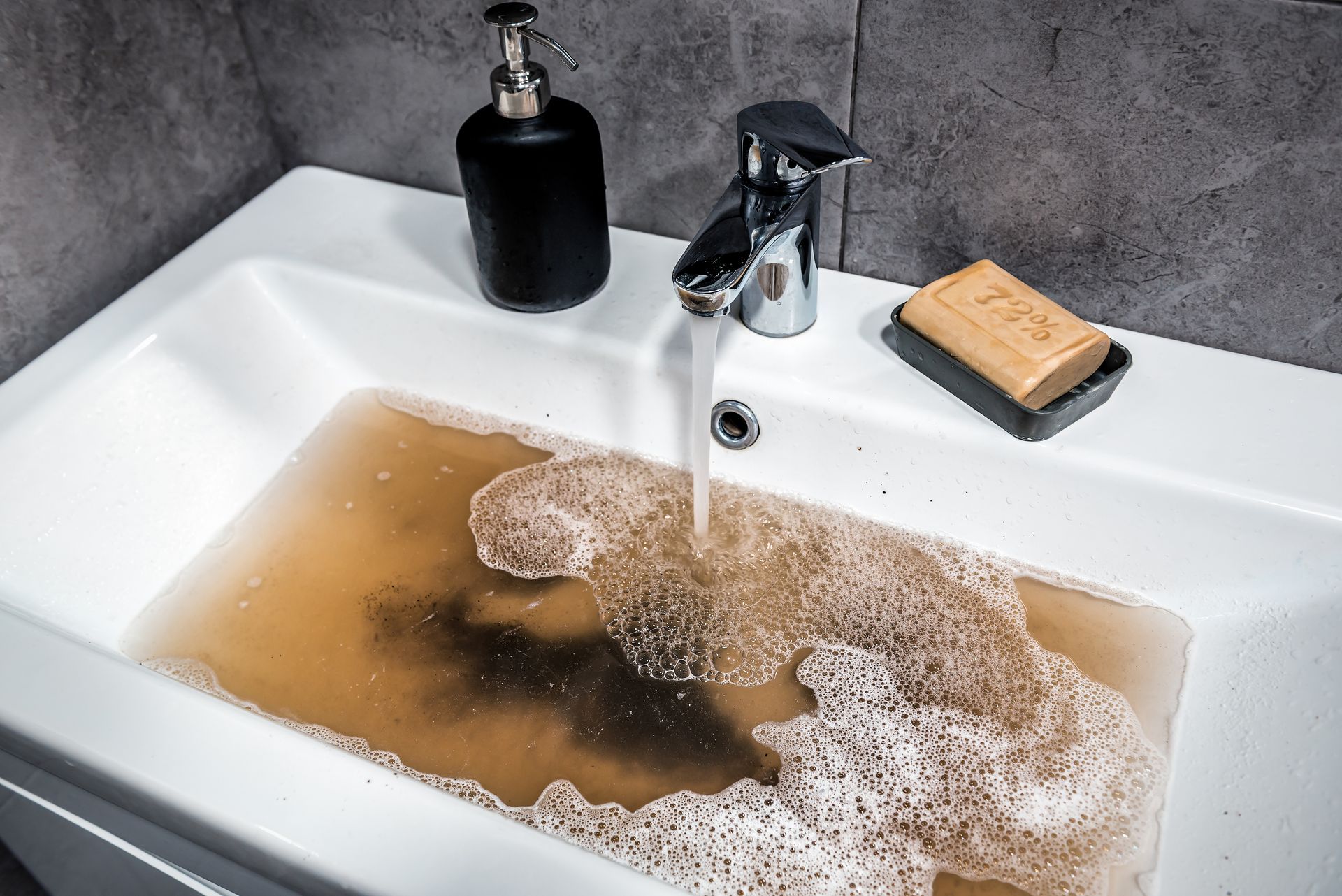 Dirty brown water overflowing in a bathroom sink with a running faucet and soap nearby