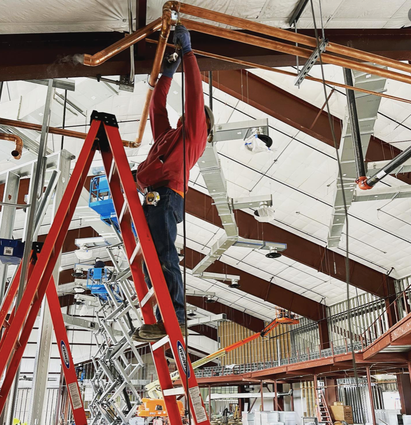 Worker on a red ladder adjusting overhead beams in a large indoor construction site