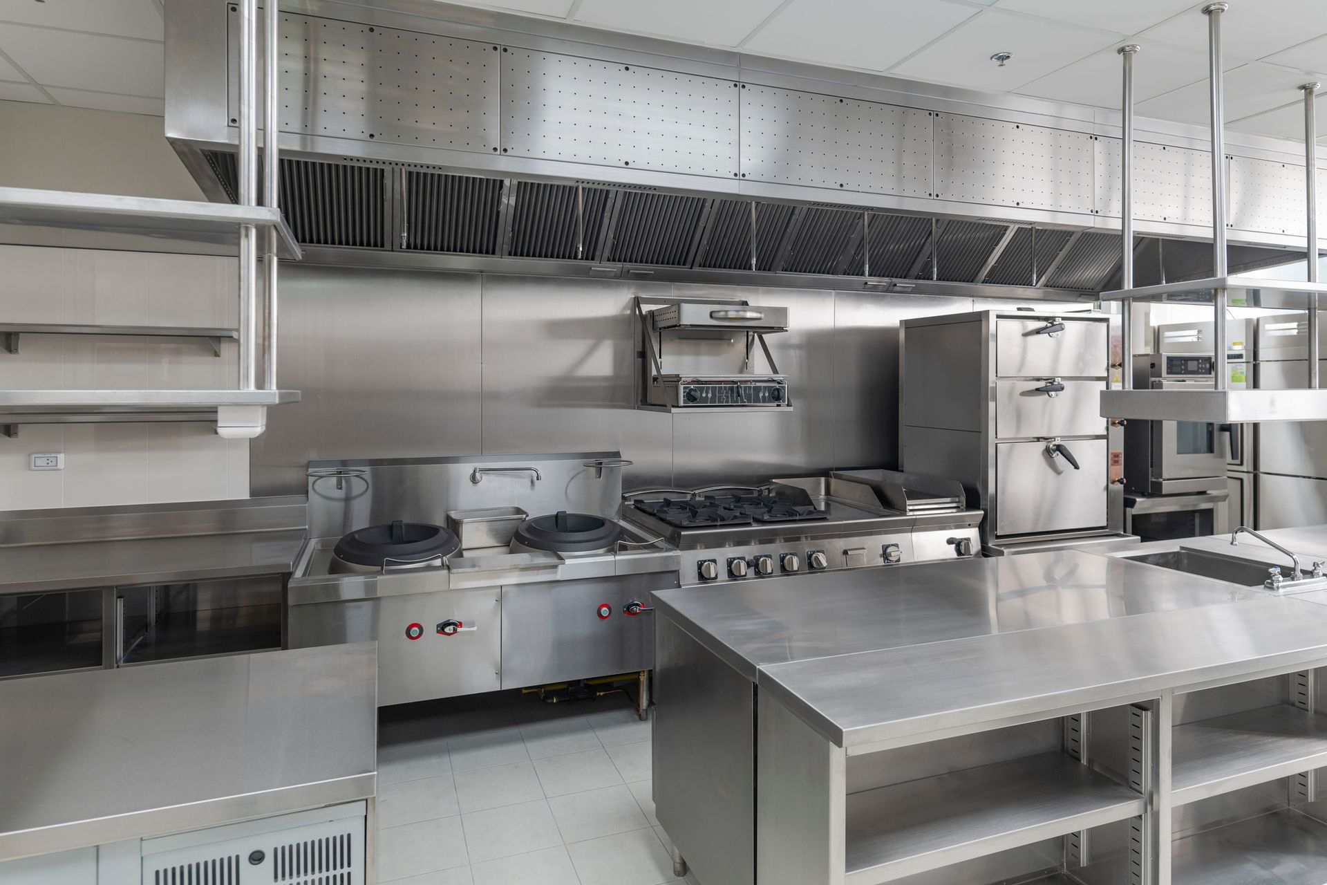 Modern stainless-steel commercial kitchen with ovens, stovetop, prep tables, and wall shelves