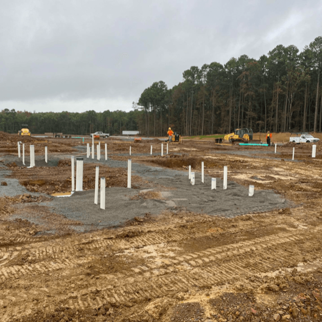 Construction site with concrete footings and white pipes in muddy ground, trees in the background