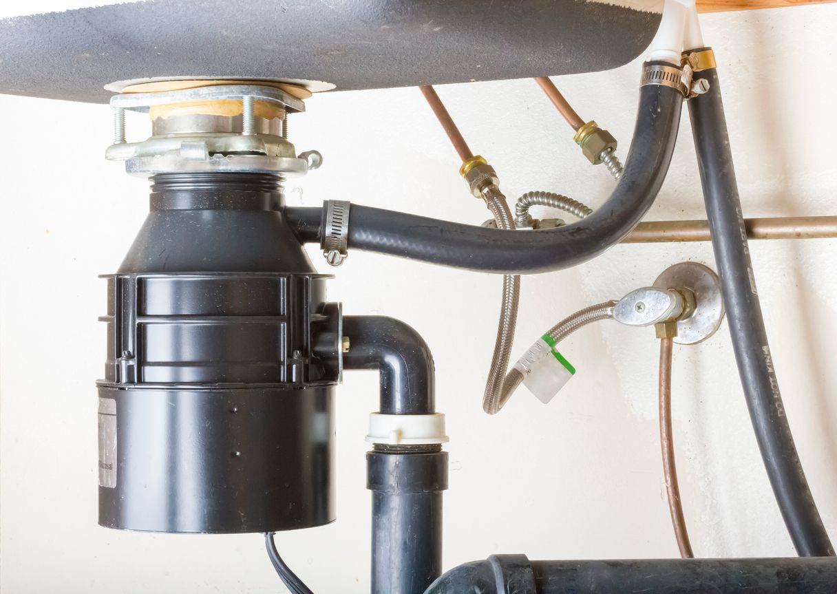 Under-sink garbage disposal with black drain pipes and hoses mounted beneath a kitchen sink
