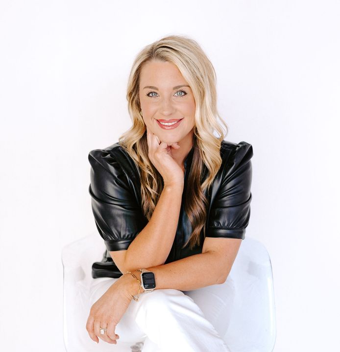 Smiling woman in black top and white pants seated against a white background