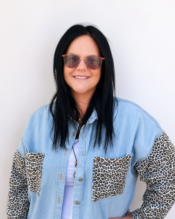 Person with long dark hair and sunglasses, wearing a light blue denim shirt with leopard-print sleeves, standing against a white wall.