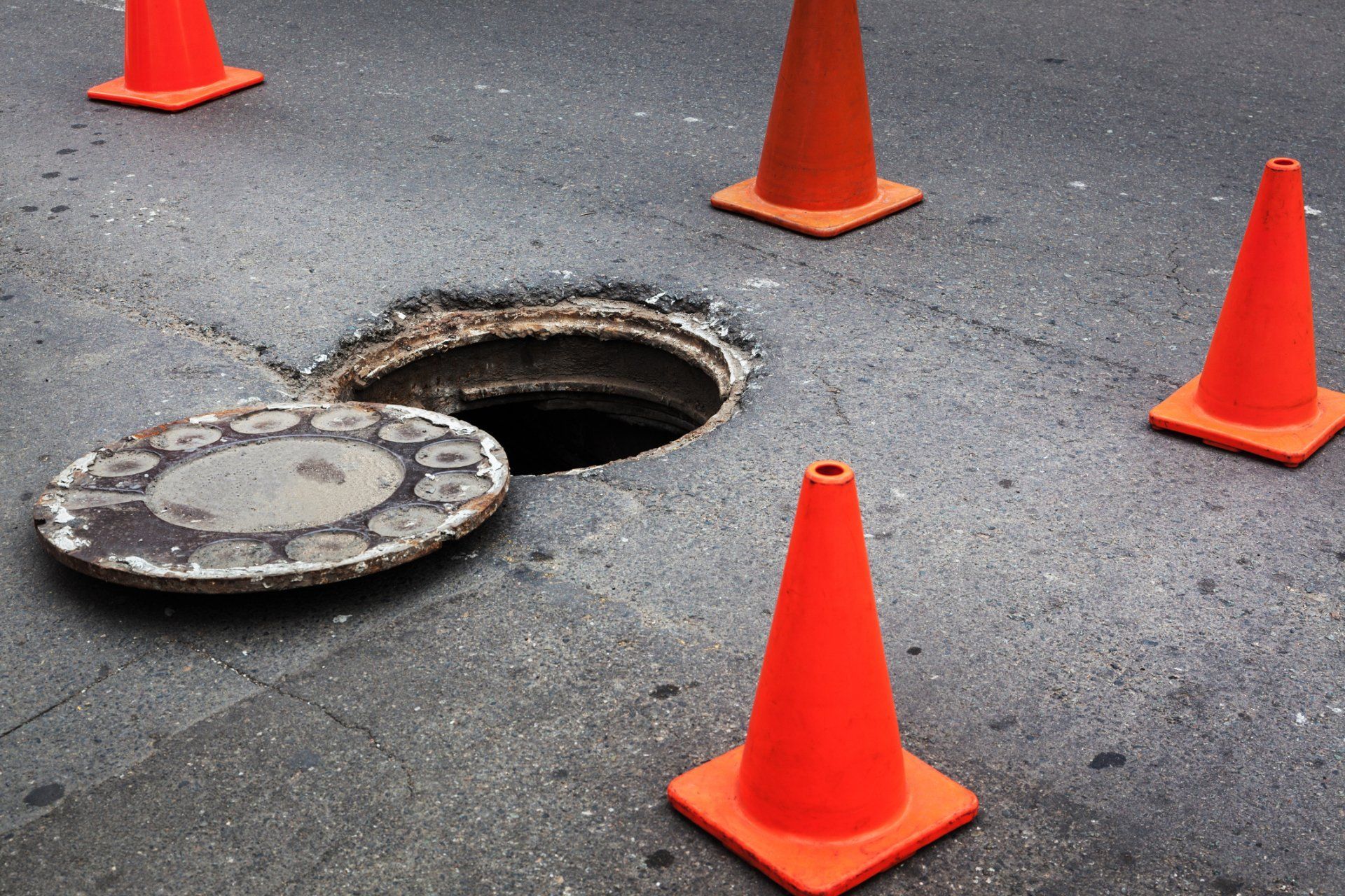 Orange cones surrounding an open manhole with the cover lying beside it on asphalt.