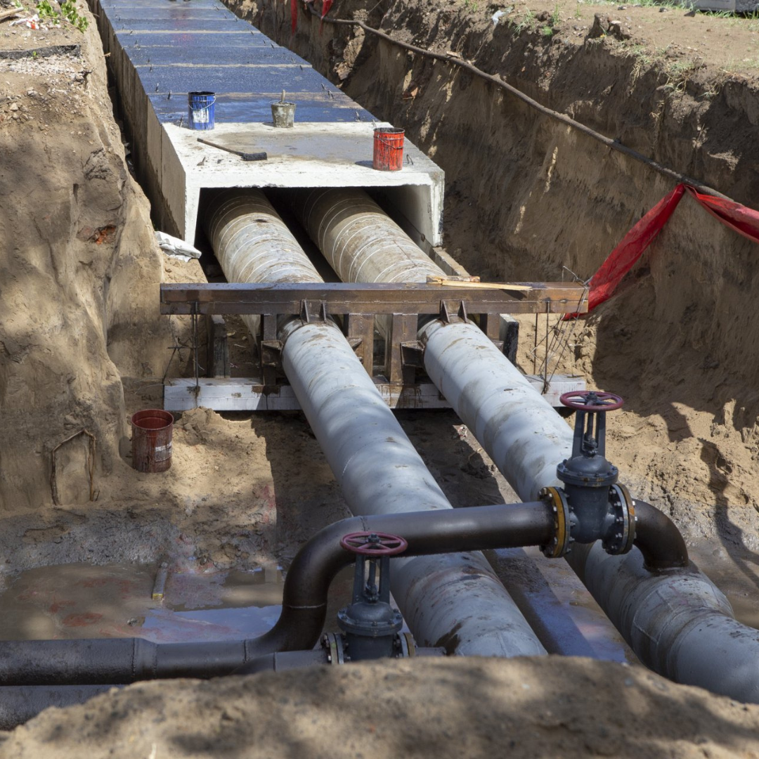Trench with exposed large gray pipes and valves beneath a concrete utility crossing.