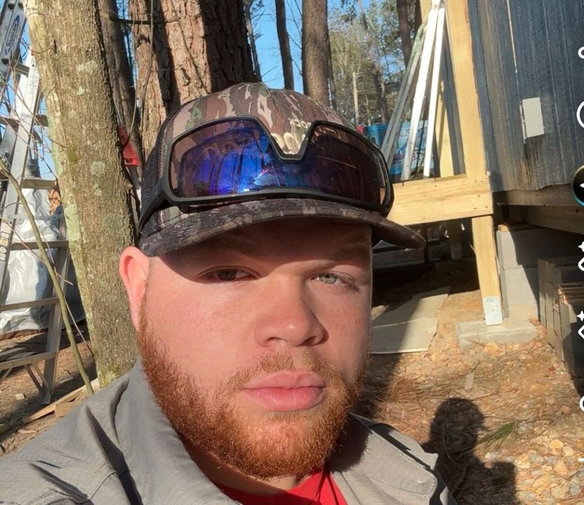 Close-up selfie of a man in a camo cap and sunglasses, outdoors by trees and a wooden structure.