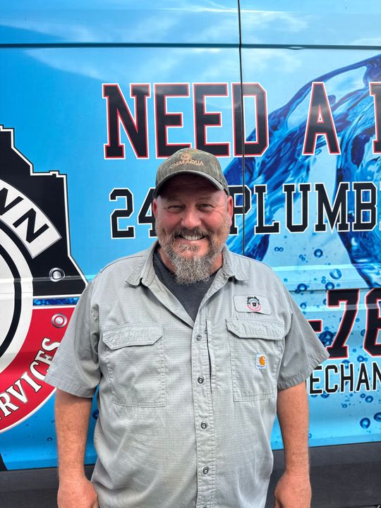 Smiling man in a cap and gray shirt stands before a blue plumbing service truck.