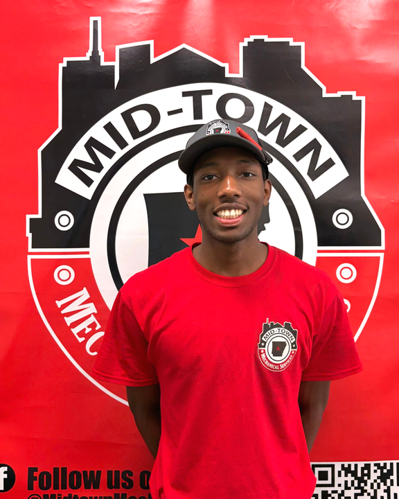 Person in red shirt and black cap standing before a Mid-Town logo backdrop