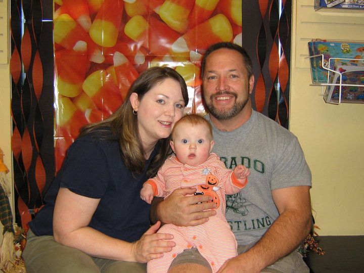 A family of three poses in front of a candy corn backdrop. A woman and man flank a baby held by the man.