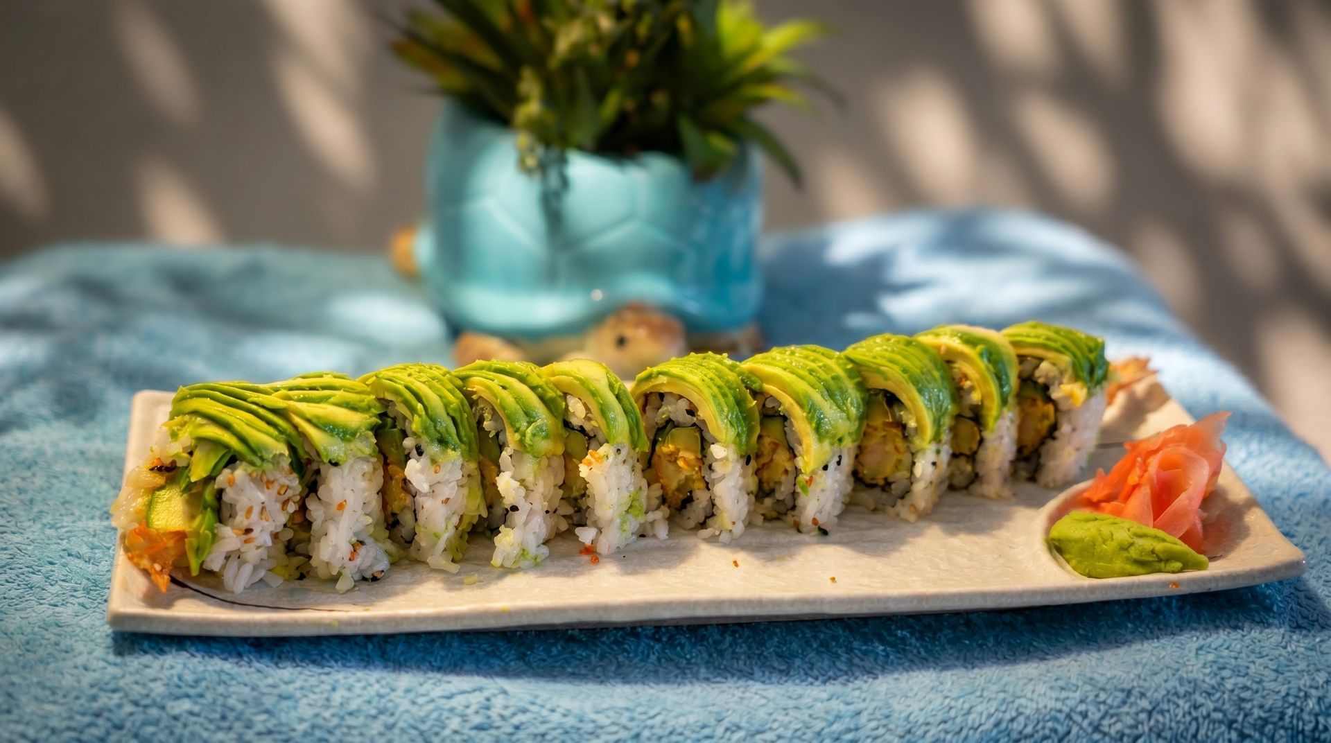 Sushi roll topped with avocado on a white plate, with ginger and wasabi on a blue tablecloth.