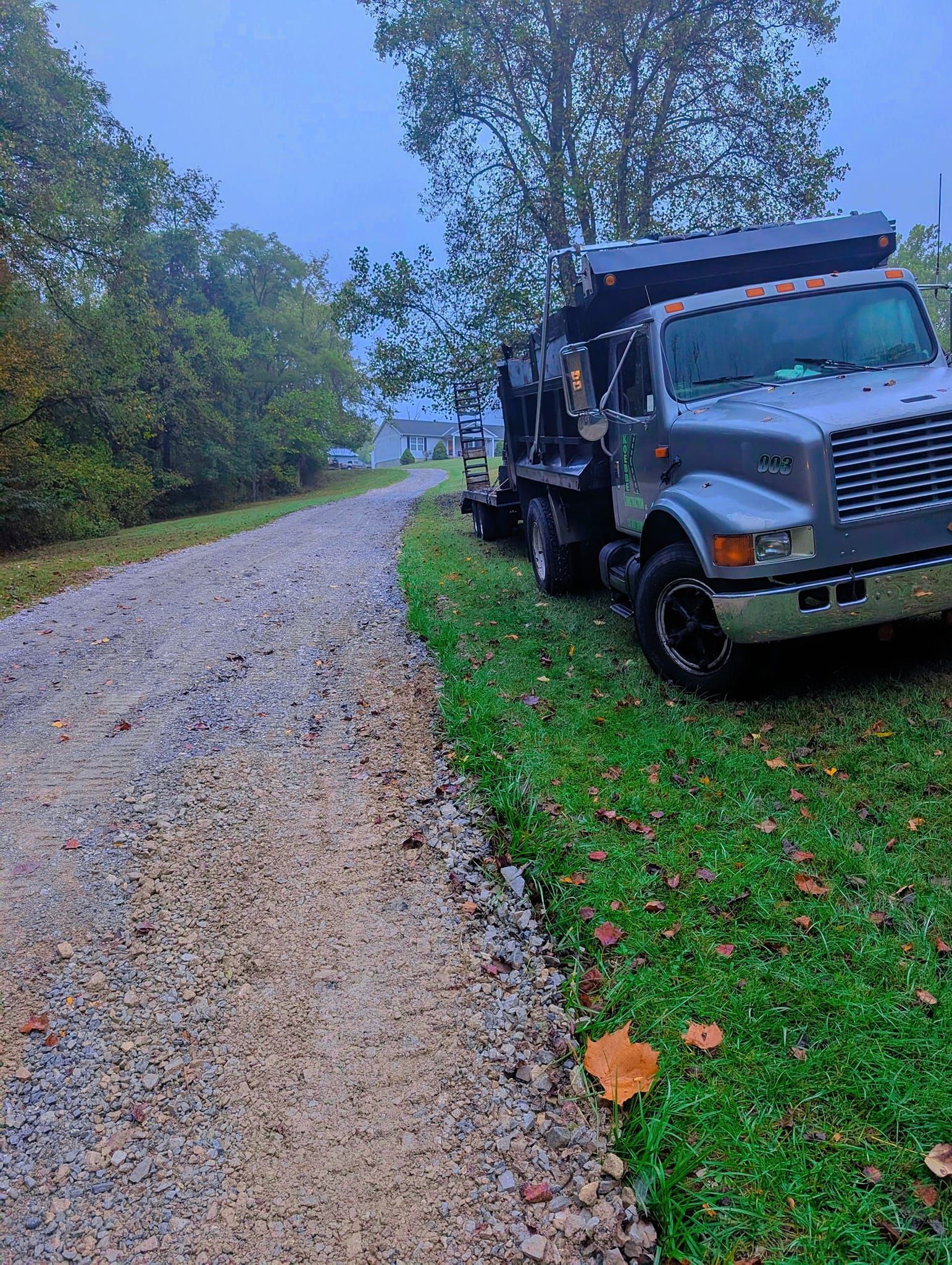 Gravel driveway next to grass with a gray dump truck parked. Overcast, trees in background.