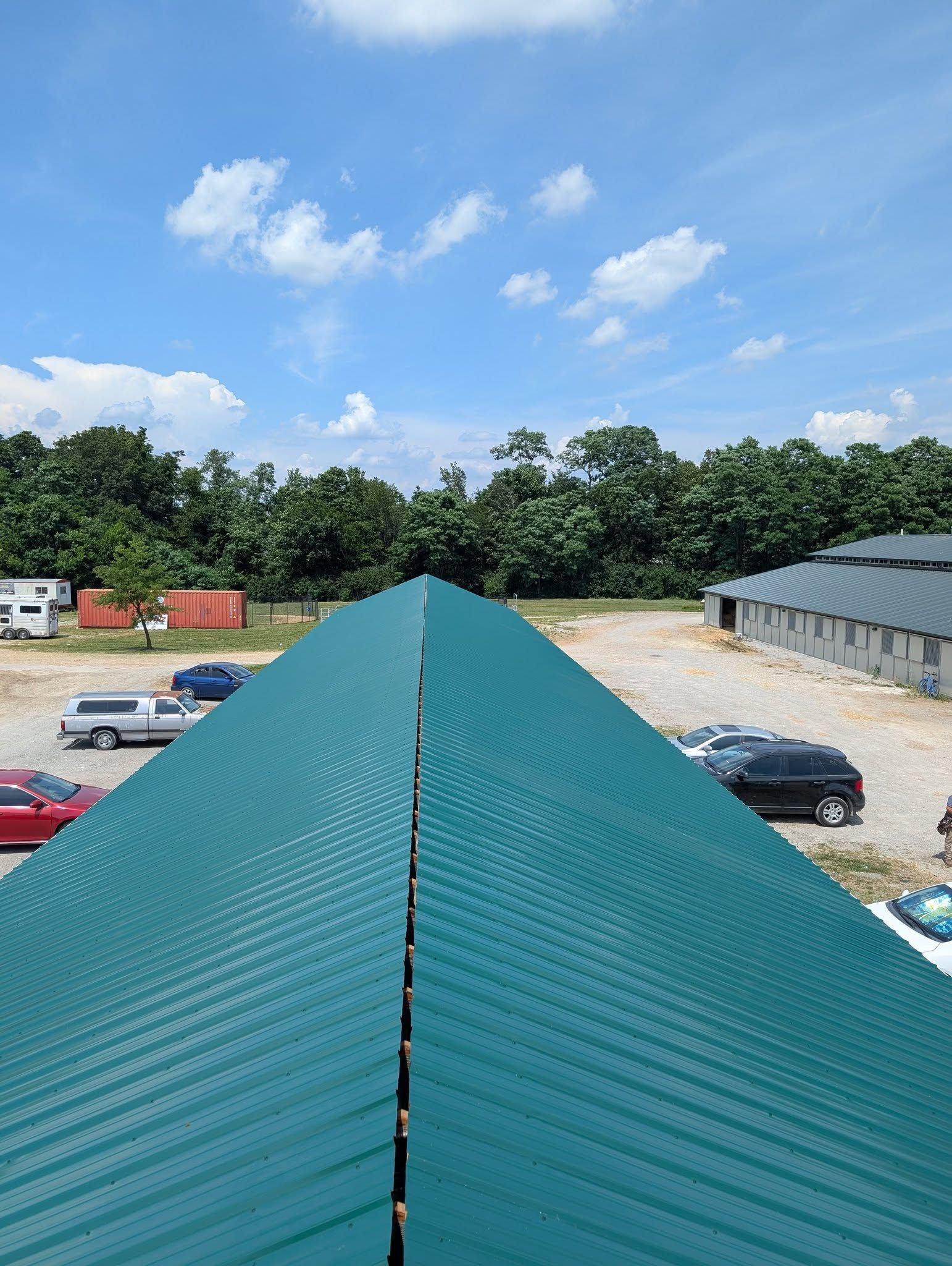 Green corrugated metal roof under blue sky with cars in parking lot and trees in the background.
