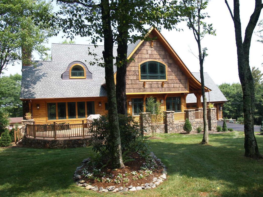 Wooden cabin-style house with a gray roof and porch, surrounded by trees and a grassy yard.