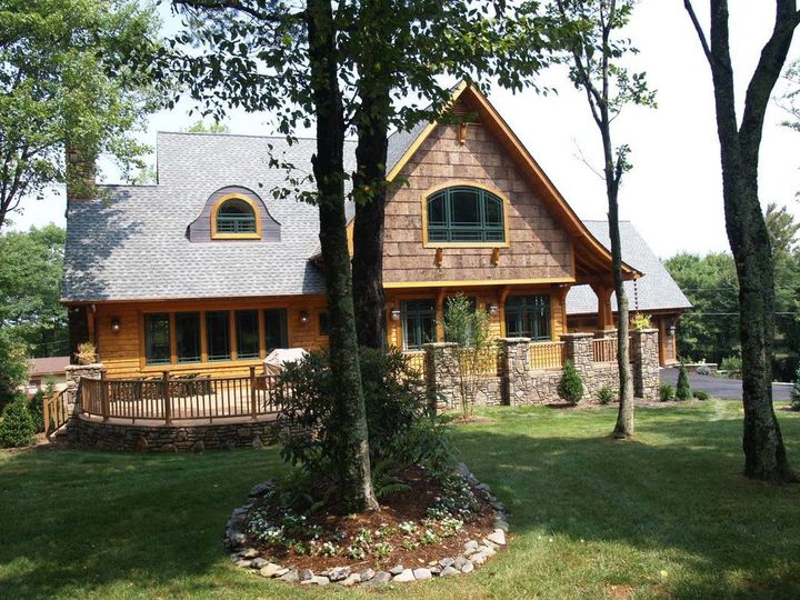 Wooden cabin-style house with a gray roof and porch, surrounded by trees and a grassy yard.