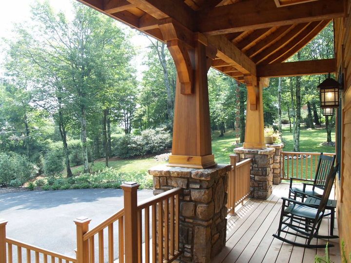 Covered wooden porch with stone columns, rocking chair, and view of a tree-lined road and grassy yard