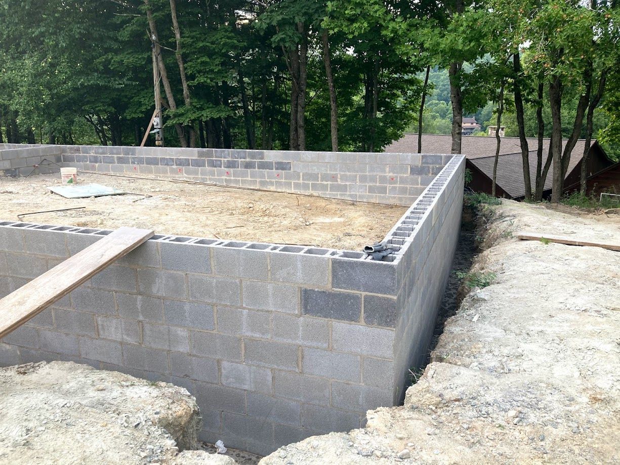 Partially built concrete foundation walls on a dirt lot surrounded by trees