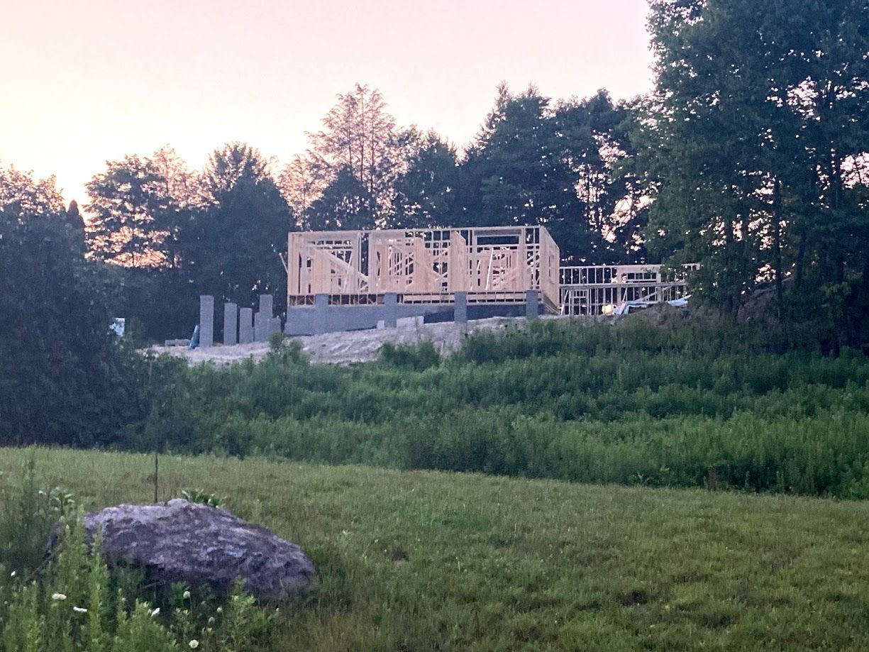 Wooden house frame under construction beyond a grassy field at dusk, with trees and a large rock in foreground