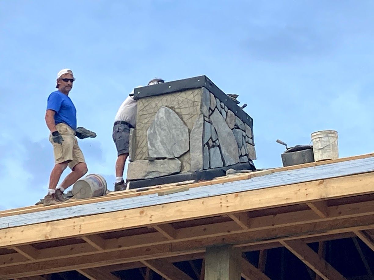 Two workers on a roof beside a stone chimney under a clear blue sky