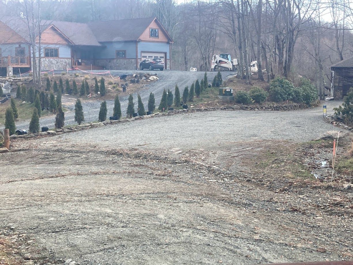 Gravel driveway leading to a house with a garage, lined with small evergreen shrubs in a wintery yard.