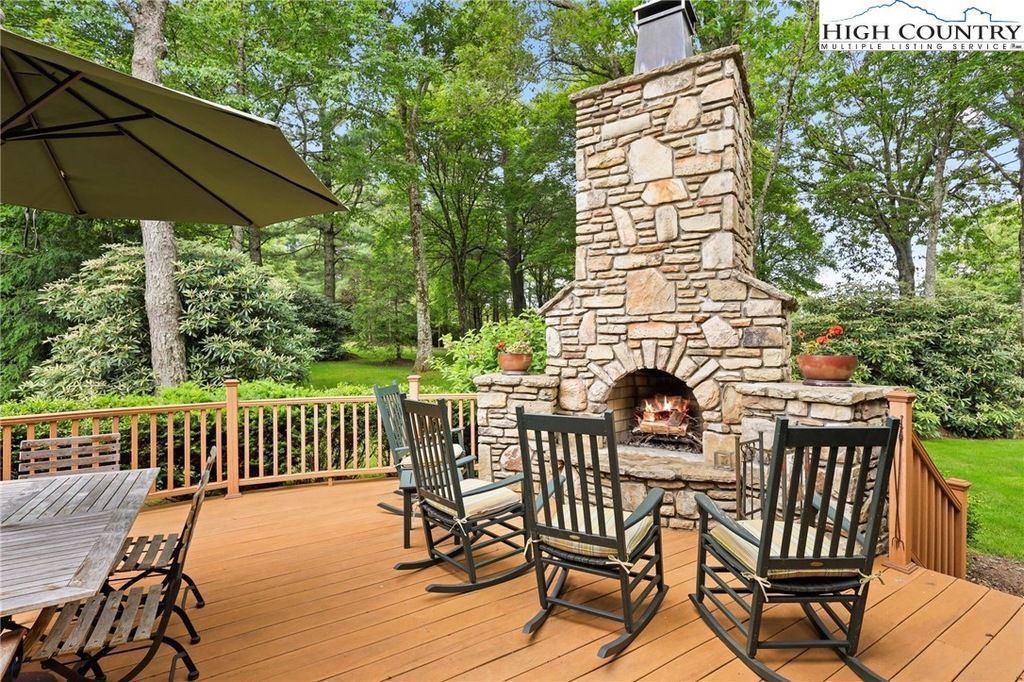 Stone outdoor fireplace on a wooden deck with black chairs, surrounded by green trees.