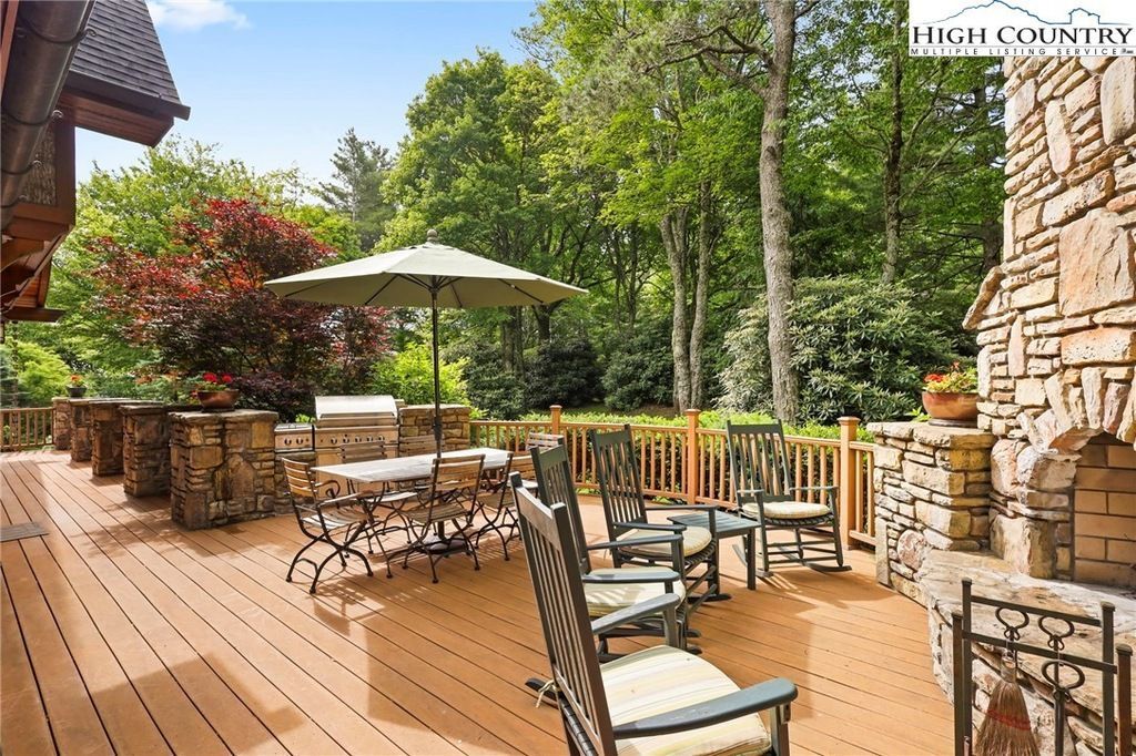 Outdoor patio with wooden deck, stone walls, tables, chairs, and a large umbrella surrounded by greenery.