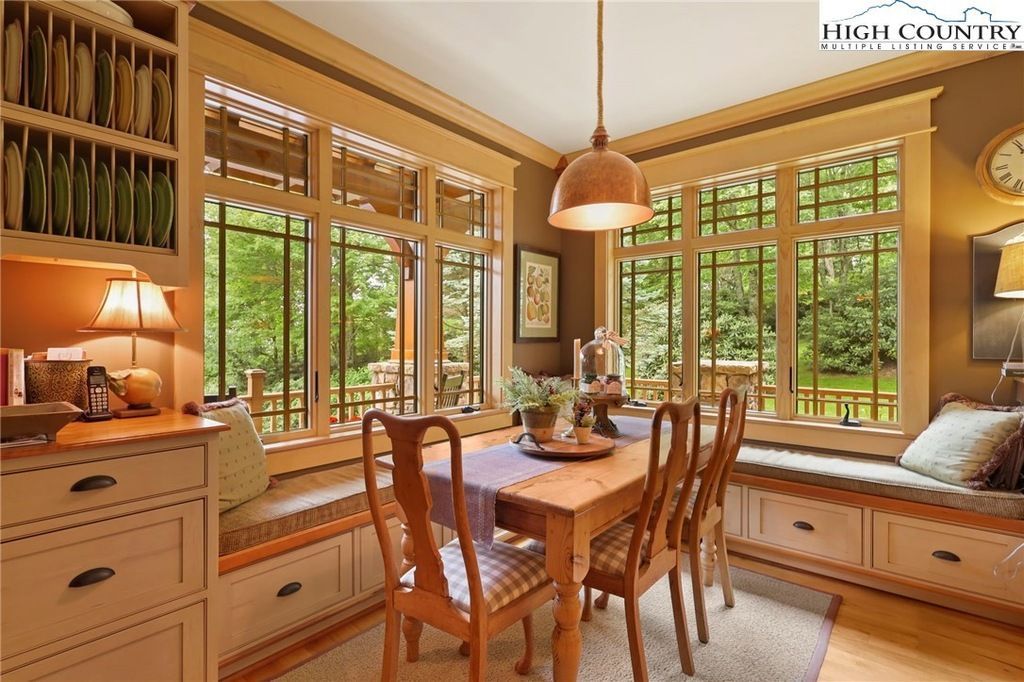 Sunlit dining nook with wooden table, chairs, and large windows overlooking greenery