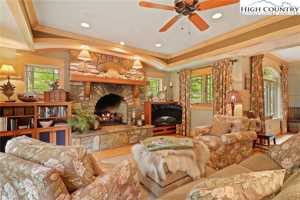 Spacious living room with stone fireplace, ornate furniture, ceiling fan, and large windows.