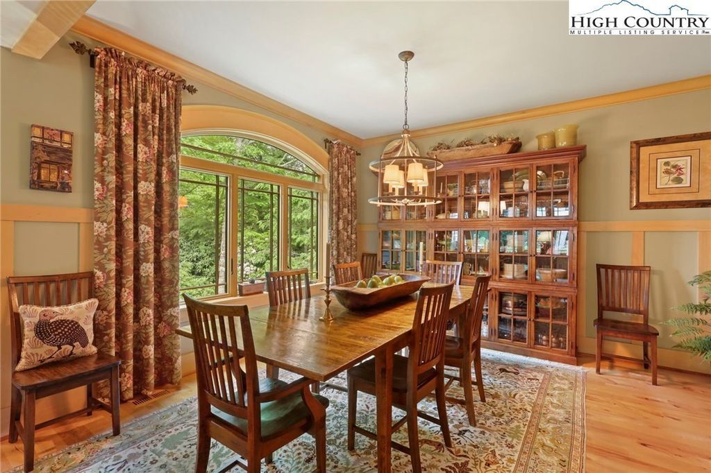 Warm dining room with wooden table, chairs, china cabinet, and large arched window overlooking greenery