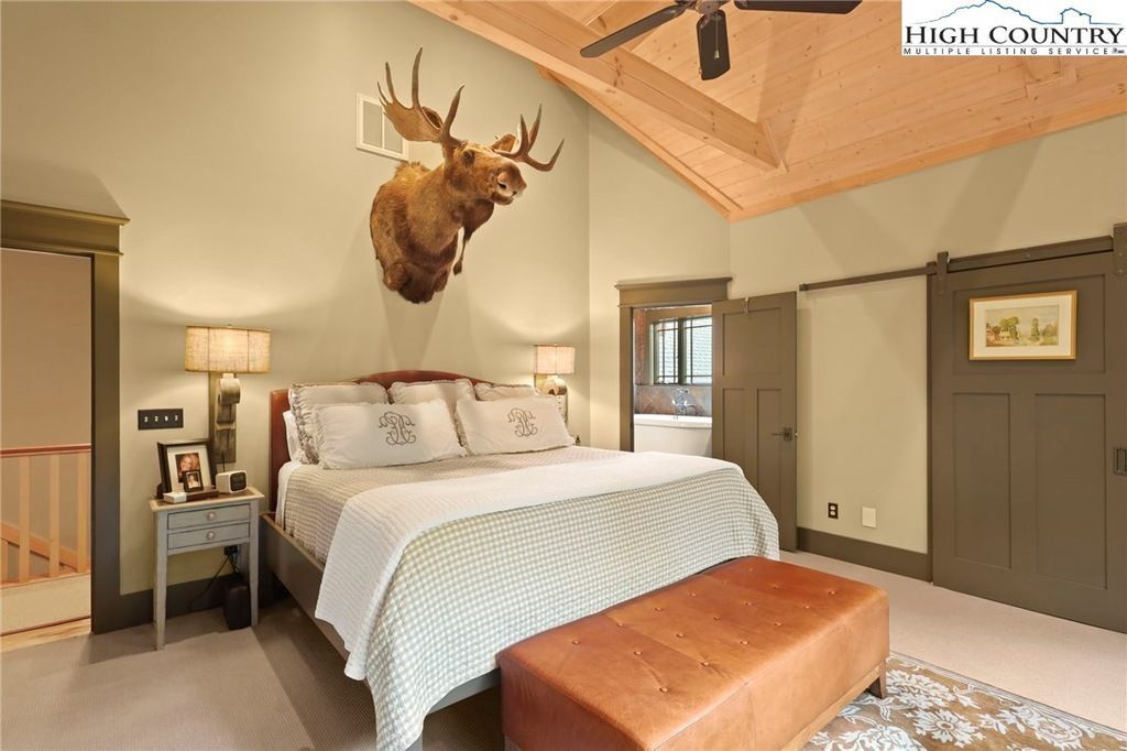 Cozy rustic bedroom with a mounted deer head, white bed, and tan bench under a vaulted ceiling