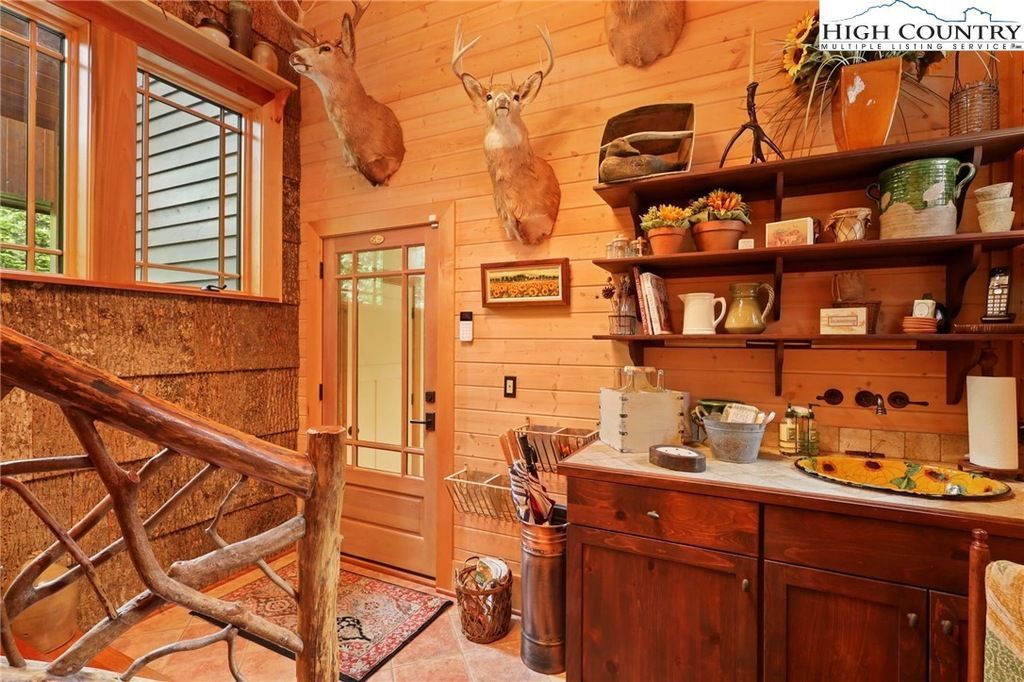 Rustic cabin interior with a wooden chair, mounted deer heads, and shelves of decor and tableware.