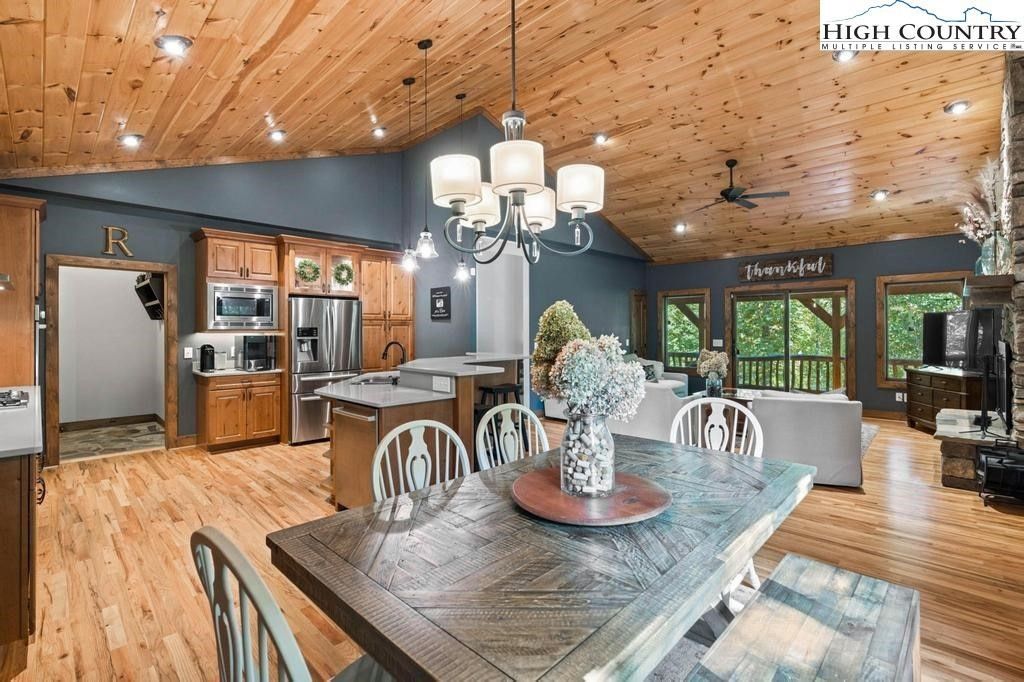 Open dining room with wood ceiling, rustic table, chandelier, and large windows overlooking greenery