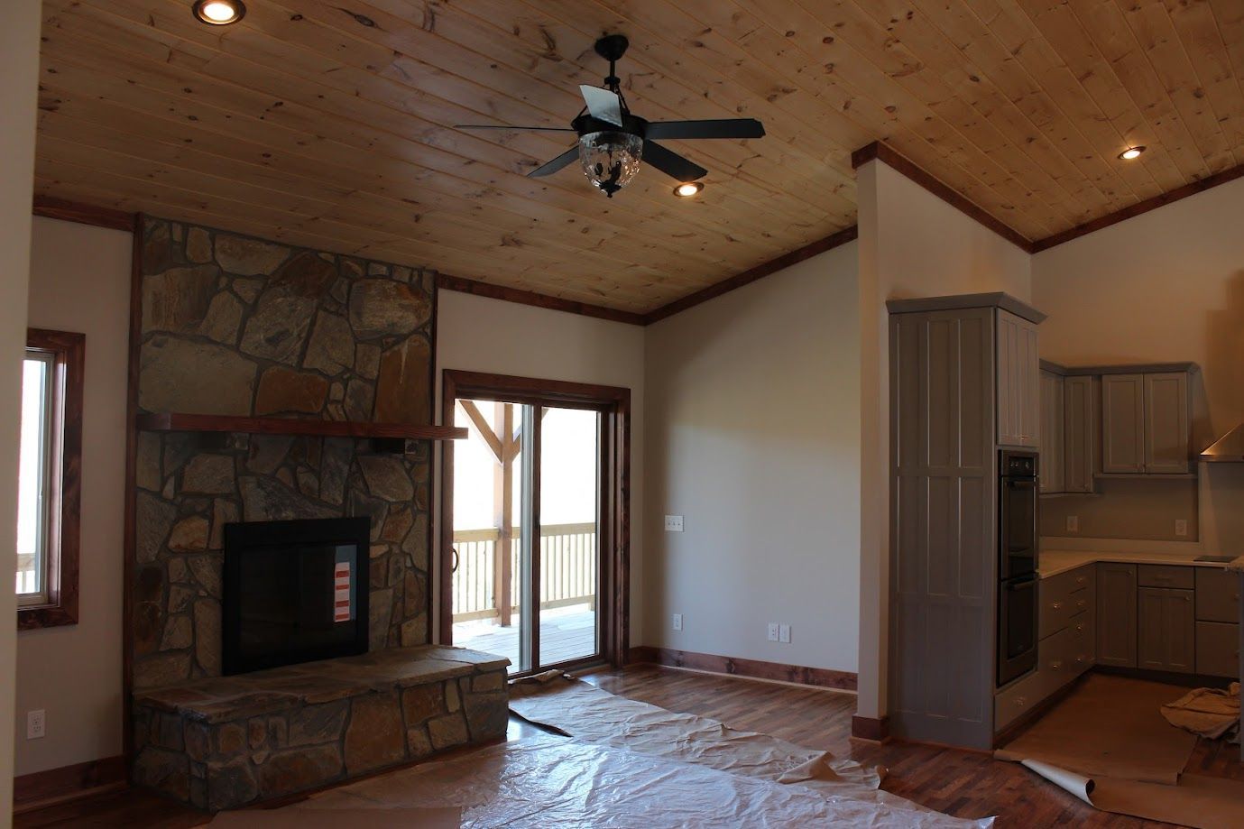 Empty living room with stone fireplace, wood ceiling, ceiling fan, and open kitchen area