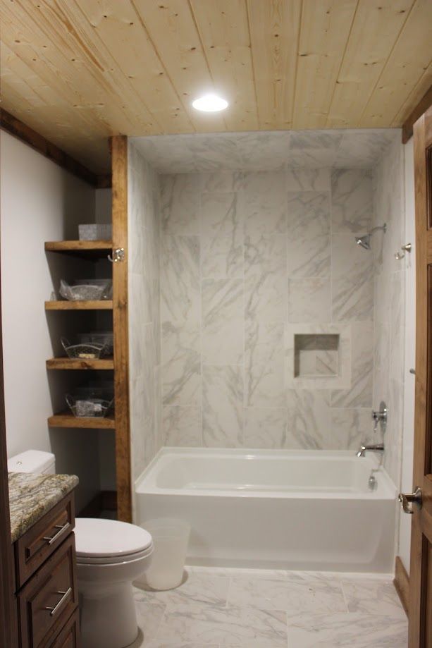 Small bathroom with a soaking tub, marble-look shower wall, wooden ceiling, and open shelving beside a toilet