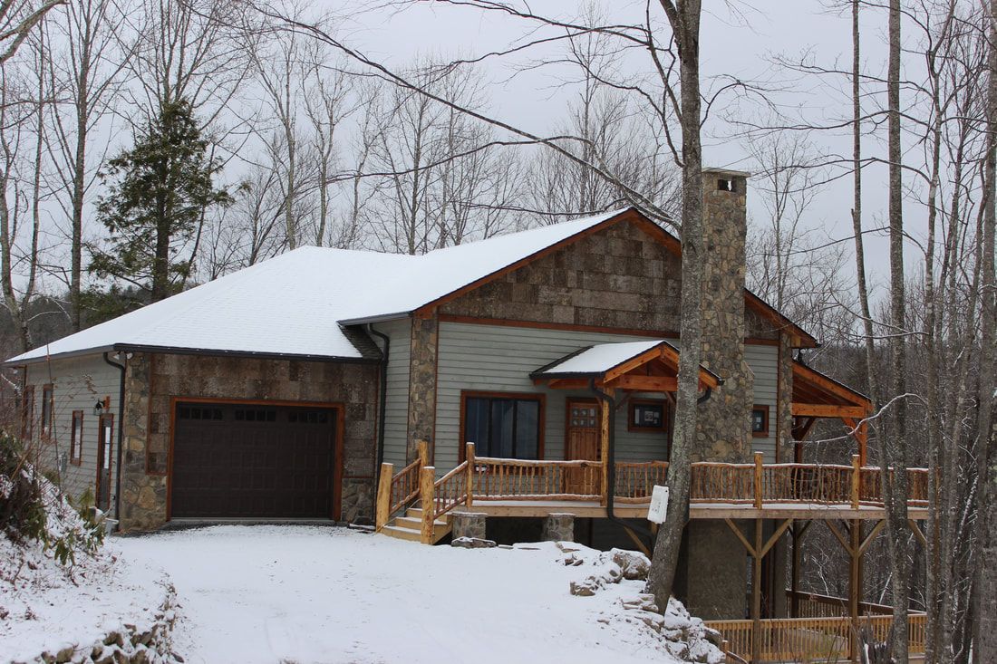 Snow-covered cabin with a garage, stone chimney, and wooden porch in a wooded winter setting